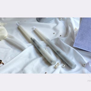 May include: Two long, cylindrical taper candles featuring a white base with grey marbled veining. The candles are positioned on a white fabric surface alongside white calla lilies, small white pearls, two gold-toned rings, and a light purple square card.