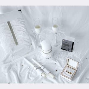 May include: A white wedding set featuring a guest book with silver trim and white feathers, two champagne flutes with feather accents, a pillar candle, two taper candles with silver bands, and an open ring box containing two gold rings.