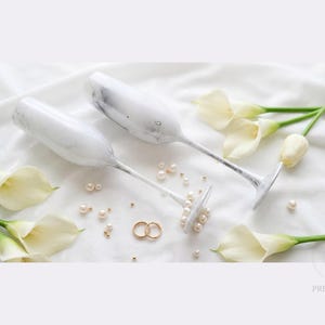 Op de afbeelding: Twee champagneglazen met een wit en grijs marmerpatroon, vergezeld van verspreide witte parels, twee gouden ringen en verschillende witte calla-lelies. De tekst "luxury PREMDECOR" is zichtbaar in de rechterbenedenhoek.