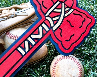 Puede incluir: Un tomahawk de madera en rojo, blanco y azul, con las letras "MM" visibles, descansa sobre hierba verde junto a una pelota de béisbol y un guante de béisbol. El tomahawk tiene un contorno azul y un diseño blanco y dorado.