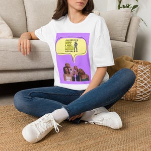 Op de afbeelding: Een wit T-shirt met korte mouwen en een rechthoekige paarse afbeelding. Het ontwerp bevat een gele tekstballon met de tekst "ALL MEN DO IS PEE LOUD AND LIE" en een kleine illustratie van een persoon, boven een foto van drie individuen.