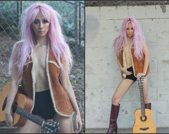 May include: A woman with pink hair wearing a brown suede vest with a white fur collar and black shorts. She is holding an acoustic guitar with a brown wooden body and a black pickguard.