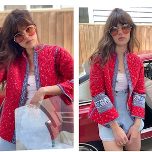 May include: A red quilted jacket with blue and white floral trim on the sleeves. The jacket has a mandarin collar and a button closure. The model is wearing the jacket with a white vest top and blue denim shorts.