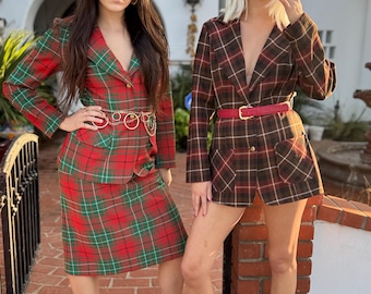 Op de afbeelding: Twee mensen in geruite blazers en rokken. De ene outfit is rood, groen en wit, de andere bruin, rood en wit. Beide outfits worden gedragen met zwarte hakken en zonnebrillen. De ene heeft een gouden riem, de andere een rode riem.