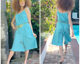May include: A light blue jumpsuit with a gold chain belt. The jumpsuit has a scoop neck and thin straps. The model is wearing the jumpsuit with a pair of gold sandals.