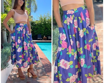 May include: A purple midi skirt with a floral print featuring pink, white, and green roses. The skirt has two large pockets on the front and a pleated waistband.