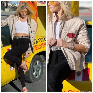 Puede incluir: Una mujer con una chaqueta beige con un parche rojo "Van Dahl", pantalones negros y un cinturón plateado. Está de pie junto a un coche amarillo con un letrero de "Entrega".