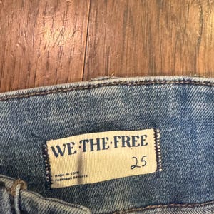 以下が含まれることがあります： 明るいブルーデニムのジーンズのクローズアップ。長方形のタグには「WE THE FREE」と書かれており、その下に数字の「25」が印刷されています。タグには「MADE IN EGYPT」と「FABRIQUE EN EGYPTE」の文字も含まれています。