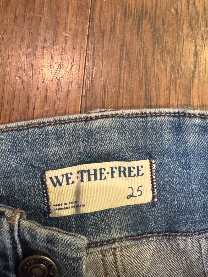 以下が含まれることがあります： 明るいブルーデニムのジーンズのクローズアップ。長方形のタグには「WE THE FREE」と書かれており、その下に数字の「25」が印刷されています。タグには「MADE IN EGYPT」と「FABRIQUE EN EGYPTE」の文字も含まれています。