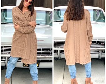 May include: A long brown cable knit cardigan with a belt. The cardigan is worn over blue jeans and brown ankle boots. The person is standing in front of a white car.