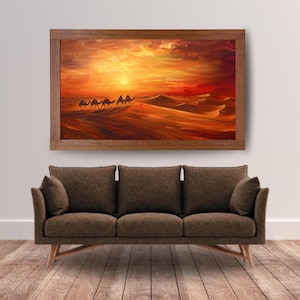 Può includere: Un'opera d'arte murale rettangolare raffigurante un paesaggio desertico con una carovana di cammelli che cammina su dune di sabbia sotto un vibrante tramonto arancione e giallo, racchiusa in una cornice di legno marrone sopra un divano in tessuto marrone.