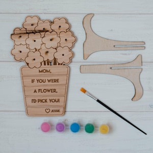Könnte beinhalten: Ein Holz-Bastelset in Blumenform mit dem Text "MOM, IF YOU WERE A FLOWER, I'D PICK YOU!" und "Jessie". Enthält Farbtöpfe, einen Pinsel und zwei Holzständer. Das Set ist für ein DIY-Projekt konzipiert.