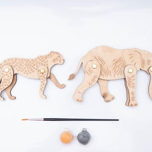 May include: Two laser-cut wooden animal figures, a cheetah and an elephant, featuring articulated limbs attached with gold-toned metal fasteners. A thin black paintbrush and two small circular paint pots in orange and gray are positioned below.