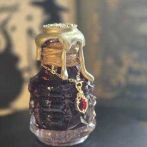 Può includere: Una piccola bottiglia di vetro sfaccettato riempita con un liquido viola scuro. Il collo è avvolto da uno spago e sigillato con cera dorata che cola. Una delicata catenina dorata pende attorno al collo con un ciondolo a pietra rossa.