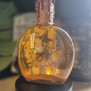 Puede incluir: Una botella de vidrio ámbar redondeada con un girasol en relieve en la parte frontal. El cuello está envuelto en cuerda y sellado con cera de color rojo oscuro. Una delicada cadena dorada con un pequeño colgante de sol cuelga del cuello.