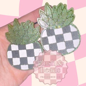 May include: Two pineapple-shaped resin accessories. The pineapples have a checkered black and white body and green glittery leaves. The text "Blush & Beads FRAGRANCE CO" is visible in the image.