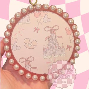 May include: A circular resin ornament featuring a light pink background with a repeating pattern of castles, bows, and mouse-ear silhouettes. The edge is decorated with a border of white pearls. The text Blush & Beads Fragrance Co is visible.