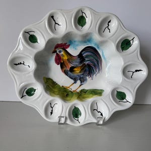 May include: A white ceramic egg serving platter with a scalloped edge featuring twelve indentations. The center is hand-painted with a colorful rooster standing on green grass against a blue sky. Each indentation is decorated with a small green leaf.