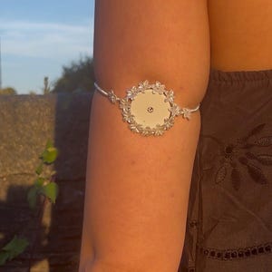 Può includere: Una copertura circolare bianca per sensore medico con una cornice decorativa color argento dal design a petali floreali. La copertura è fissata a una sottile fascia regolabile color argento che avvolge la parte superiore del braccio.