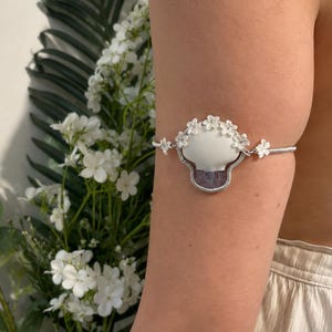 May include: An arm cuff featuring a central mushroom-shaped charm with a white cap and a purple base, wrapped in silver-toned wire. The charm is accented with small white flower embellishments along the top and sides, connected to a coiled wire band.