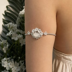 Pode incluir: Um bracelete em tom prateado com um medalhão circular central rodeado por pequenas pétalas de flores esculpidas. A banda consiste num design de fio flexível enrolado com dois pequenos detalhes florais adicionais em cada lado do centro.