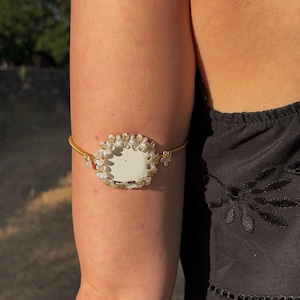 May include: An upper arm bracelet featuring a thin gold-toned metallic band. The centrepiece is a rectangular white plaque framed by a decorative border of small, white, five-petaled floral shapes that extend slightly along the band on each side.
