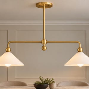 May include: A linear pendant light fixture featuring a brushed brass finish with a central vertical rod and a horizontal bar supporting two conical white glass shades. The fixture includes a circular ceiling canopy and spherical accents.