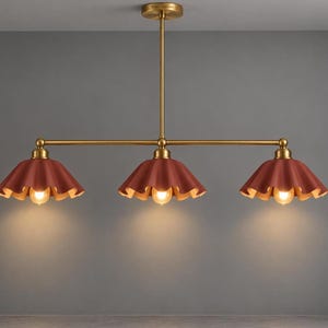 May include: A linear chandelier featuring a brass-toned metal frame and three downward-facing, ruffled-edge shades in a deep terracotta red color. Each shade houses a single glowing light bulb, suspended from a central vertical rod.