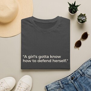 May include: A dark gray t-shirt featuring the white text "A girl's gotta know how to defend herself." The shirt is displayed alongside a straw hat, a pair of sunglasses, blue denim jeans, and white sneakers, with two small potted succulents nearby.