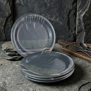 May include: A stack of four round ceramic plates featuring a dark gray glaze with a subtle, lighter gray textured pattern of vertical lines and horizontal waves across the surface. The plates have a smooth, glossy finish and a circular shape.