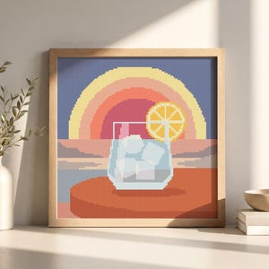 Peut inclure: Une broderie au point de croix encadrée représentant une illustration pixelisée d'un verre rempli de glaçons et d'une tranche de citron. L'arrière-plan montre un coucher de soleil avec des arcs concentriques jaunes, orange et roses.
