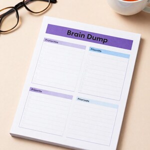 May include: A white notepad with the words "Brain Dump" in black and a purple banner. The notepad is divided into four sections labeled "Priorities" and "Priorities" in purple, and "Priorities" and "Priorities" in light blue. A pair of glasses and a cup of tea are in the background.
