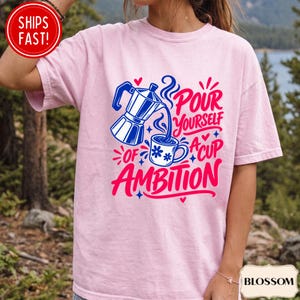May include: A light pink short-sleeve t-shirt featuring a blue moka pot pouring coffee into a blue mug with flower accents. The graphic includes the text "Pour yourself a cup of ambition" in bold pink and blue lettering with small heart and star icons.