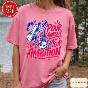 May include: A pink short-sleeve t-shirt featuring a graphic of a blue moka pot pouring coffee into a white mug with blue flowers. The text reads, "POUR YOURSELF A CUP OF AMBITION" in bold pink and blue lettering, accented by small hearts and stars.