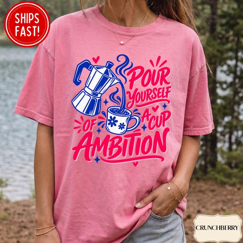 May include: A pink short-sleeve t-shirt featuring a graphic of a blue moka pot pouring coffee into a white mug with blue flowers. The text reads, "POUR YOURSELF A CUP OF AMBITION" in bold pink and blue lettering, accented by small hearts and stars.