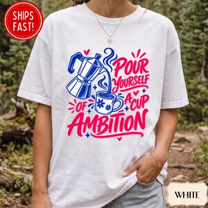 May include: A white short-sleeve t-shirt featuring a graphic of a blue moka pot pouring coffee into a mug. The design includes the text "POUR YOURSELF A CUP OF AMBITION" in bold pink and blue lettering, accented with small hearts and star shapes.