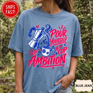 May include: A blue short-sleeve t-shirt featuring a graphic of a moka pot pouring coffee into a mug with blue flower accents. The text reads Pour Yourself A Cup Of Ambition in pink and blue script. The design includes small red hearts and blue stars.
