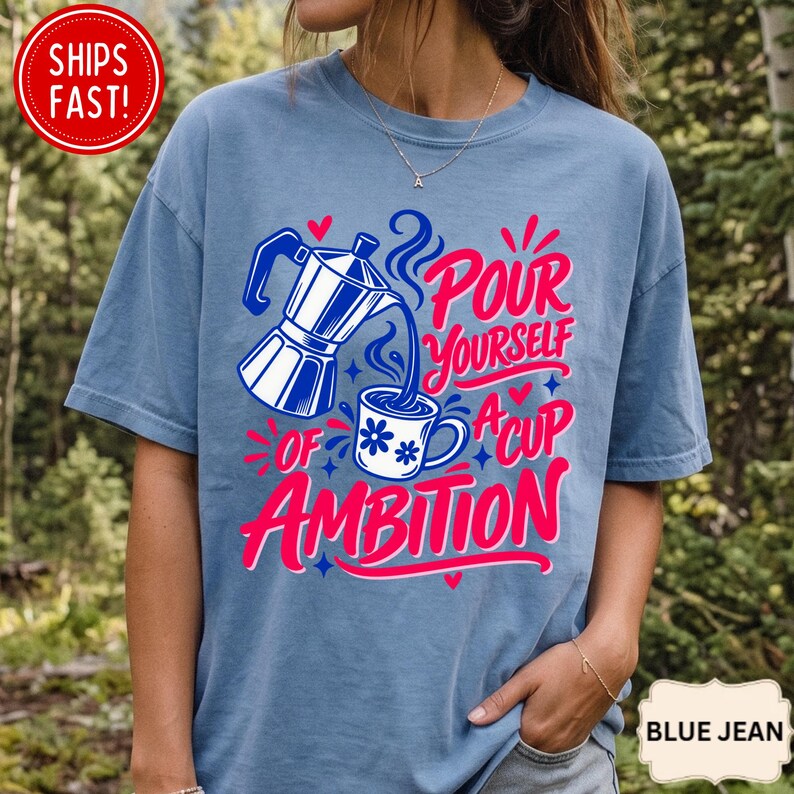 May include: A blue short-sleeve t-shirt featuring a graphic of a moka pot pouring coffee into a mug with blue flower accents. The text reads Pour Yourself A Cup Of Ambition in pink and blue script. The design includes small red hearts and blue stars.