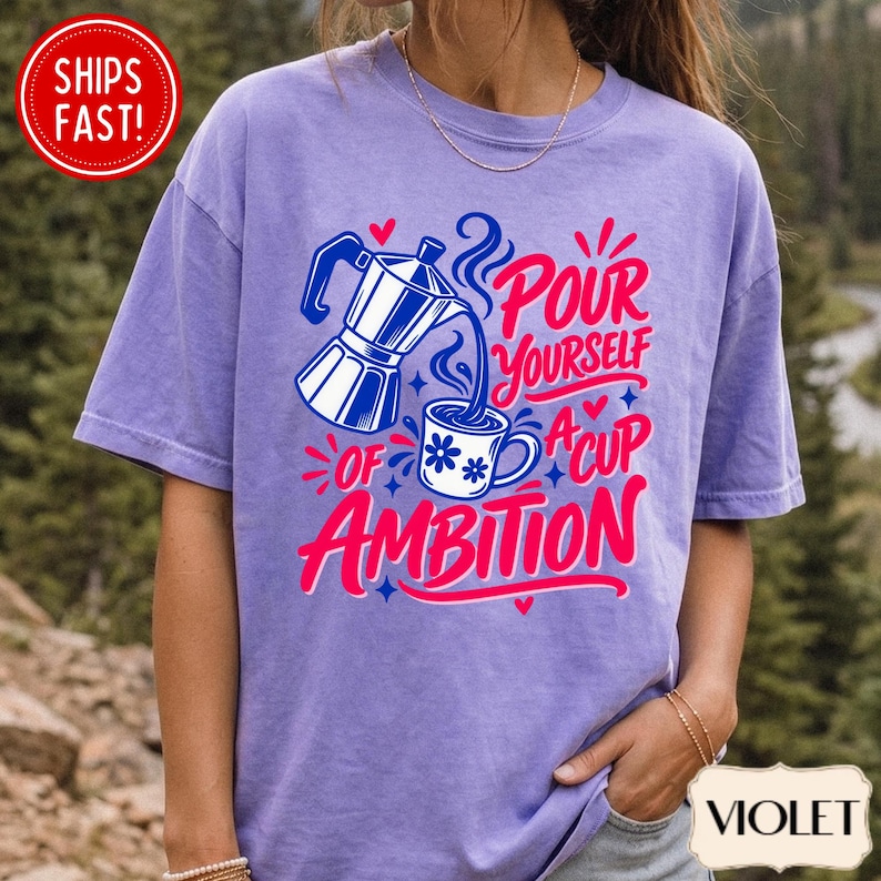 May include: A violet short-sleeve t-shirt featuring a graphic of a moka pot pouring coffee into a mug with a flower pattern. The text reads "POUR YOURSELF A CUP OF AMBITION" in pink and blue lettering, accented by small hearts and star shapes.