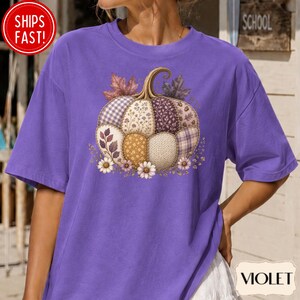 May include: A violet short-sleeved t-shirt featuring a central graphic of a pumpkin composed of various patchwork patterns, including floral, plaid, and knit textures. The pumpkin is accented with small white daisy flowers and autumn leaves.