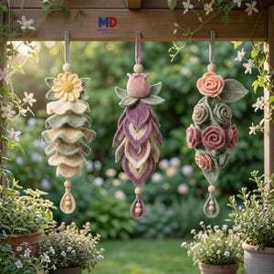 May include: Three hanging crochet ornaments featuring floral designs. The left ornament has a yellow flower and cream and sage green ruffled layers. The center ornament features a pink tulip and purple leaves. The right ornament displays pink roses and sage green foliage.