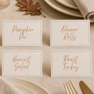 Op de afbeelding: Vier rechthoekige witte naamkaartjes met dunne randen en koperkleurige cursieve tekst met de woorden Pumpkin Pie, Dinner Rolls, Harvest Salad en Roast Turkey, gerangschikt op een tafelsetting met witte pompoenen en goudkleurig bestek.