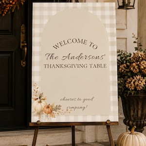 Może przedstawiać: Prostokątna tablica powitalna na drewnianej sztaludze z beżowym łukiem na jasnoszaro-białym wzorze w kratę. Tekst brzmi: WELCOME TO The Andersons' THANKSGIVING TABLE oraz cheeres to good company! z grafiką przedstawiającą dynie.