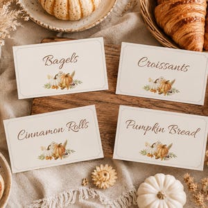May include: Four rectangular white food labels with brown cursive text reading Bagels, Croissants, Cinnamon Rolls, and Pumpkin Bread. Each label features a small illustration of pumpkins and dried foliage at the bottom center.