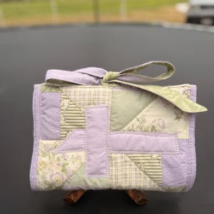 May include: A rectangular quilted fabric roll featuring a patchwork design in shades of lavender, sage green, and cream. The exterior displays various floral and plaid patterns, secured at the top with a matching fabric tie tied into a bow.
