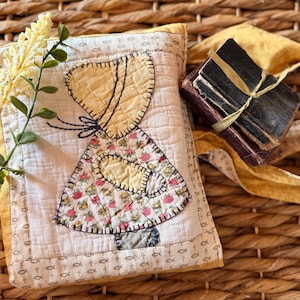 May include: A rectangular quilted fabric pouch featuring an appliqué of a figure in a bonnet and floral dress. The pouch has a white center with a fish and cross pattern border in mustard yellow. A small stack of dark books is tied with a gold ribbon.