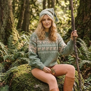 Puede incluir: Una mujer con un suéter verde con un patrón floral rosa, un gorro a juego y leggings color melocotón. Sostiene un palo de madera y está sentada en una roca cubierta de musgo en un bosque. También lleva botas marrones.