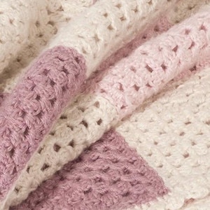 Puede incluir: Una manta de ganchillo con rayas alternas de hilo color crema, rosa claro y malva polvoriento. El tejido presenta un patrón de puntos calados consistente con una apariencia suave y texturizada, mostrando los intrincados bucles del hilo.