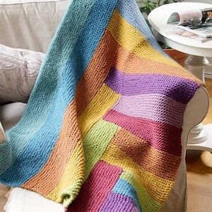 Puede incluir: Una manta tejida que presenta un vibrante patrón geométrico multicolor de bloques rectangulares en tonos azul, amarillo, naranja, verde y morado. El tejido tiene una textura de punto bobo visible en toda la superficie de la pieza.