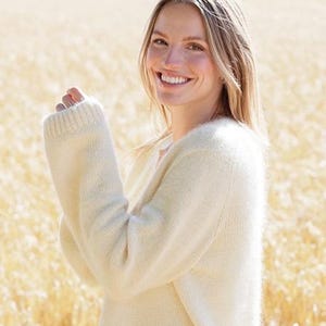 Puede incluir: Una mujer con un suéter color crema de manga larga. El suéter tiene una textura suave y difusa. El fondo es un campo de trigo dorado.